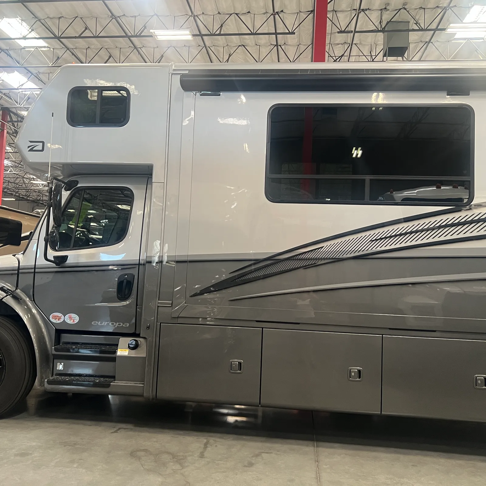 2027 Dynamax Corp Europa S 37CA Super C motorhome, side profile of motorhome with cab, windows, and storage compartments in warehouse