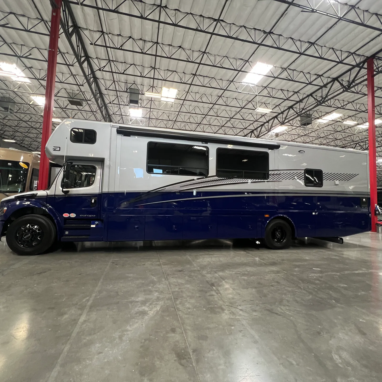 2027 Dynamax Corp Europa S 37CA Super C motorhome, motorhome with white upper and blue lower body in warehouse setting