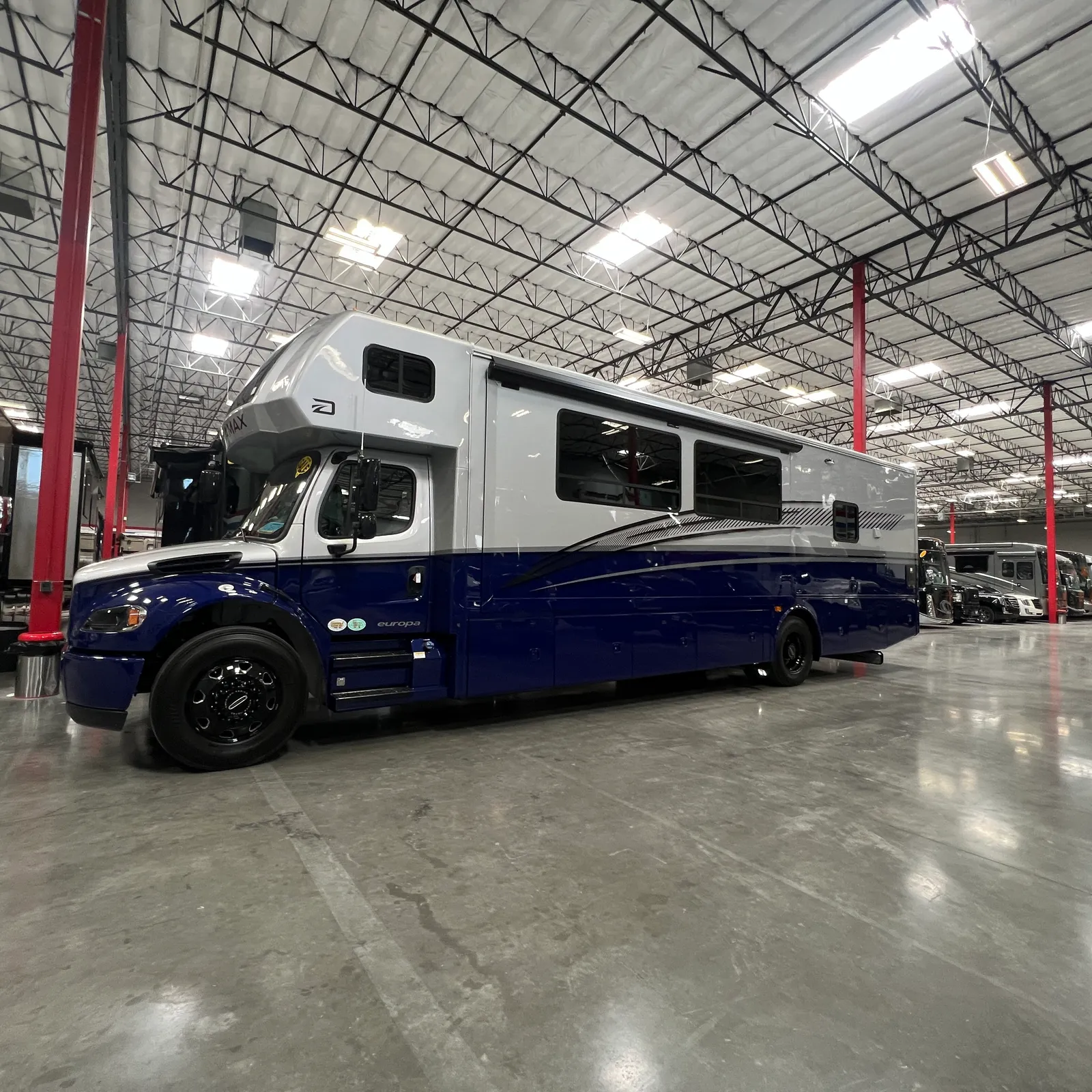 2027 Dynamax Corp Europa S 37CA Super C motorhome, white and blue motorhome in warehouse with metal support structure