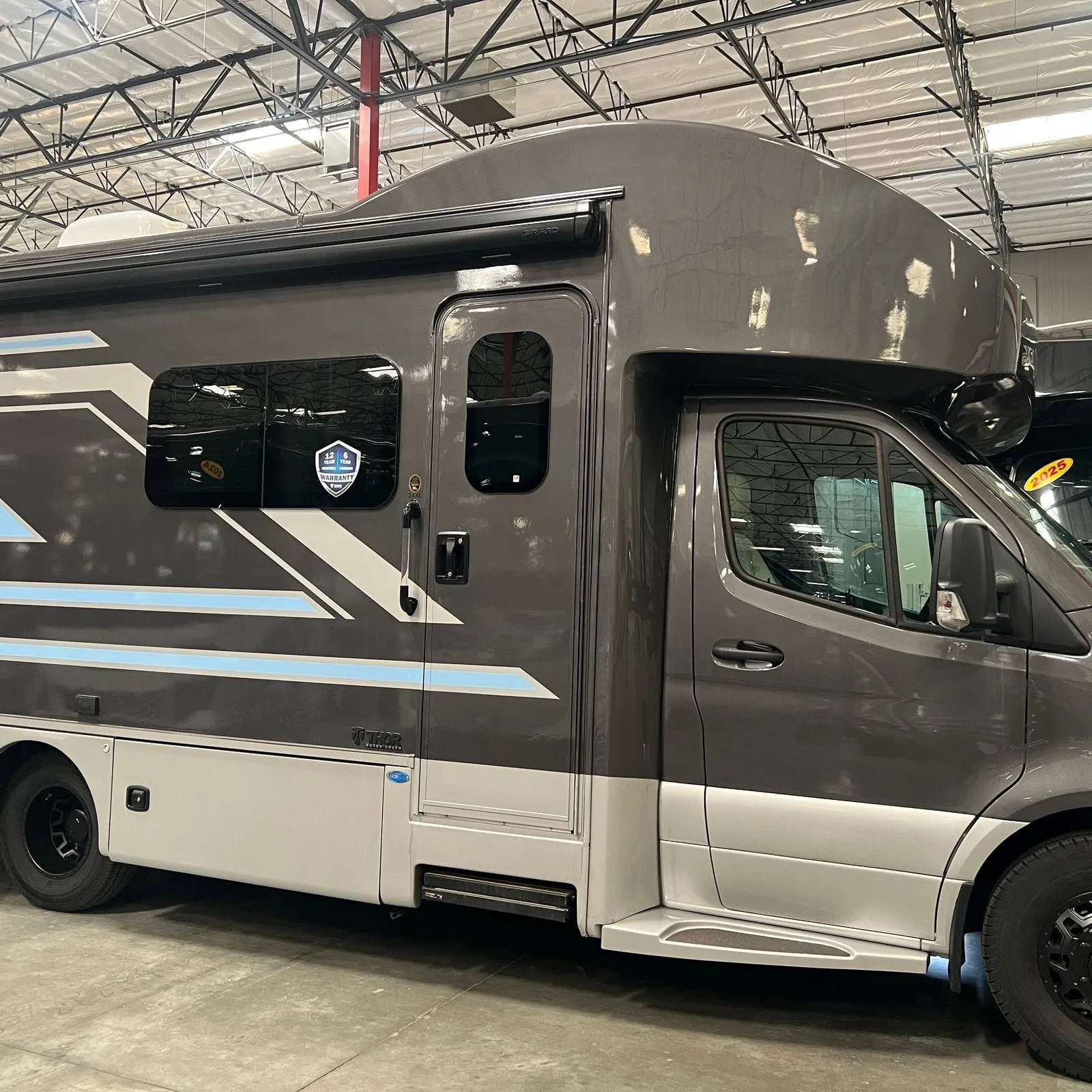 2026 Thor Delano 24XL Class C motorhome, gray and white motorhome with blue stripe detail, parked indoors